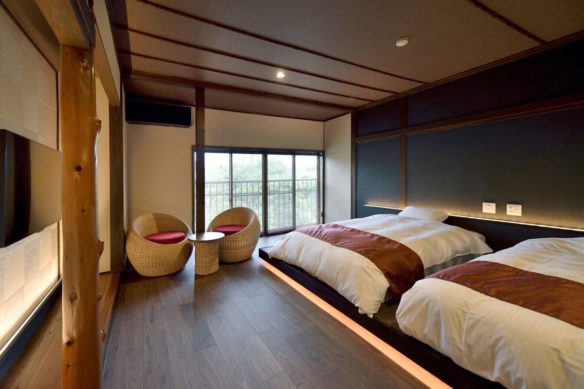 Une chambre avec deux lits, un plancher en bois, une grande fenêtre et des chaises en osier.