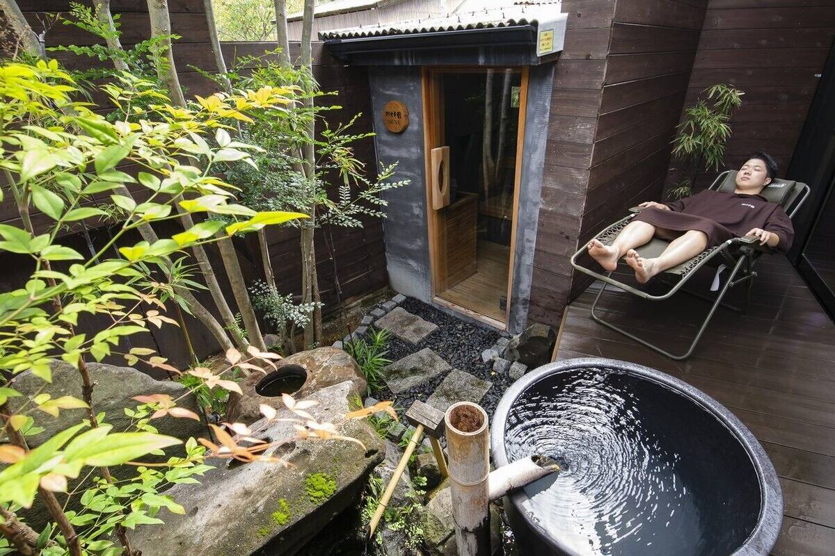 Une personne se détend sur un fauteuil dans un espace aménagé en bois, agrémenté d’une fontaine en pierre et de verdure.