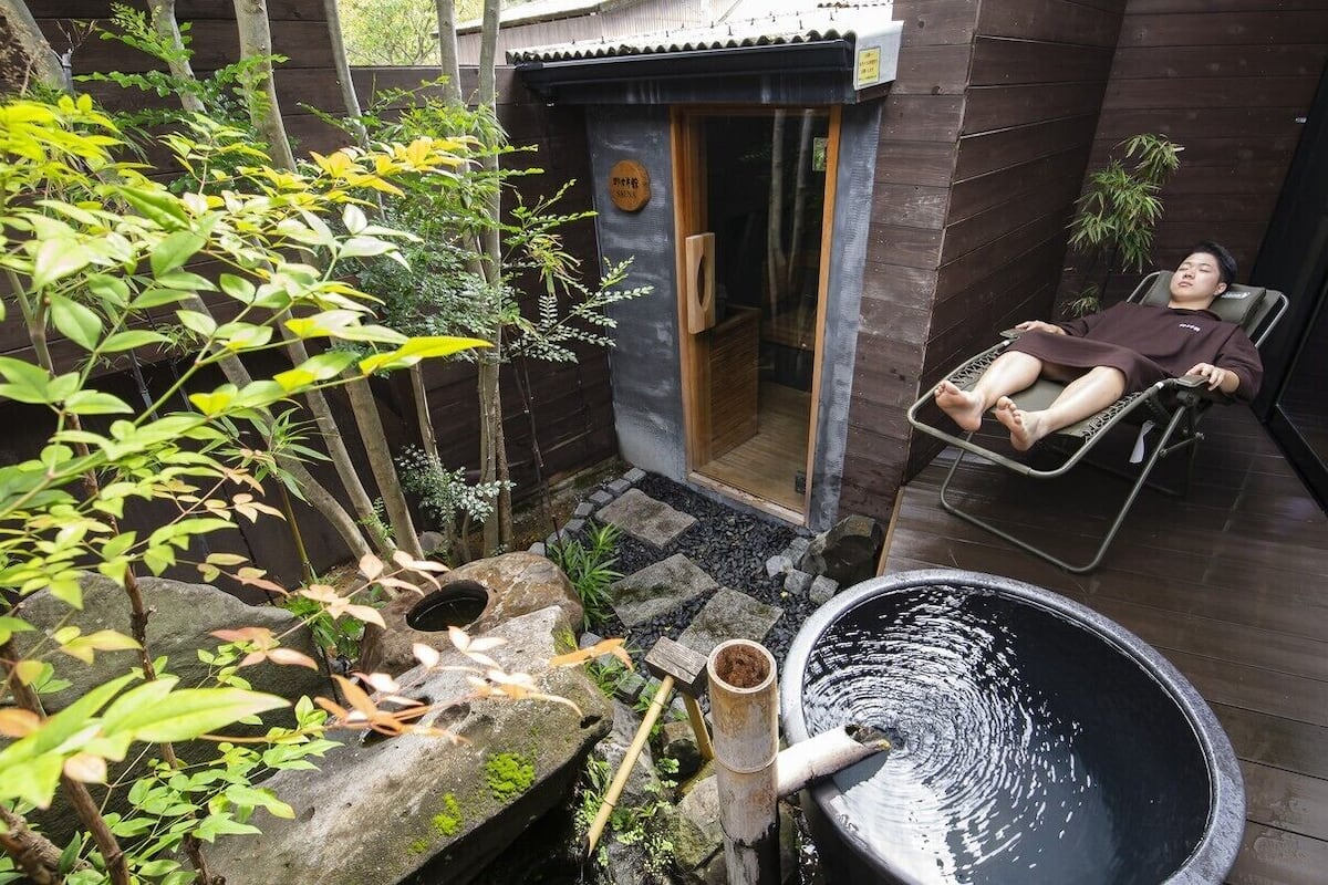 Iemand ontspant in een loungestoel op een houten terras, met een waterpartij van natuursteen en groen.