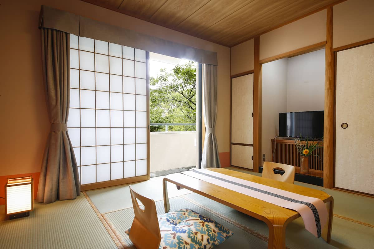 A traditional Japanese room with tatami flooring, a low wooden table, a television, and a sliding door leading to a balcony with a view of greenery.