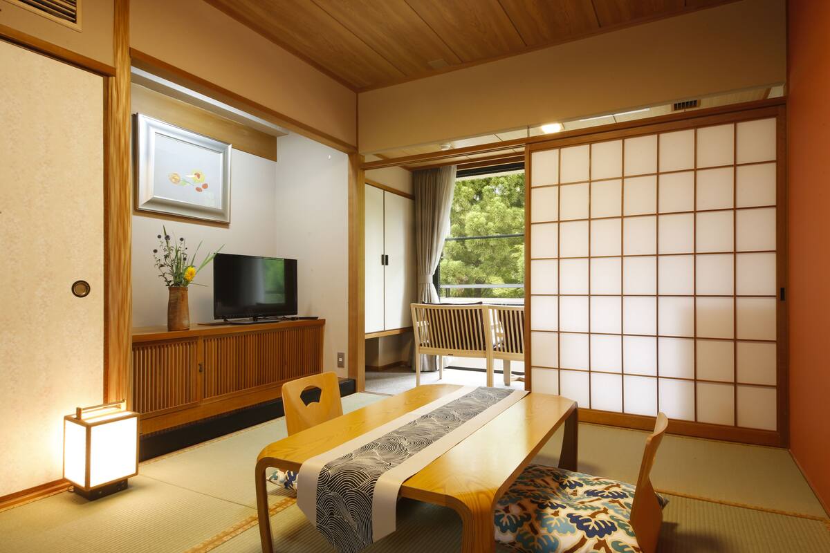 A traditional Japanese room with tatami flooring, a low table with a table runner, a television on a wooden cabinet, and a sliding shoji screen door.