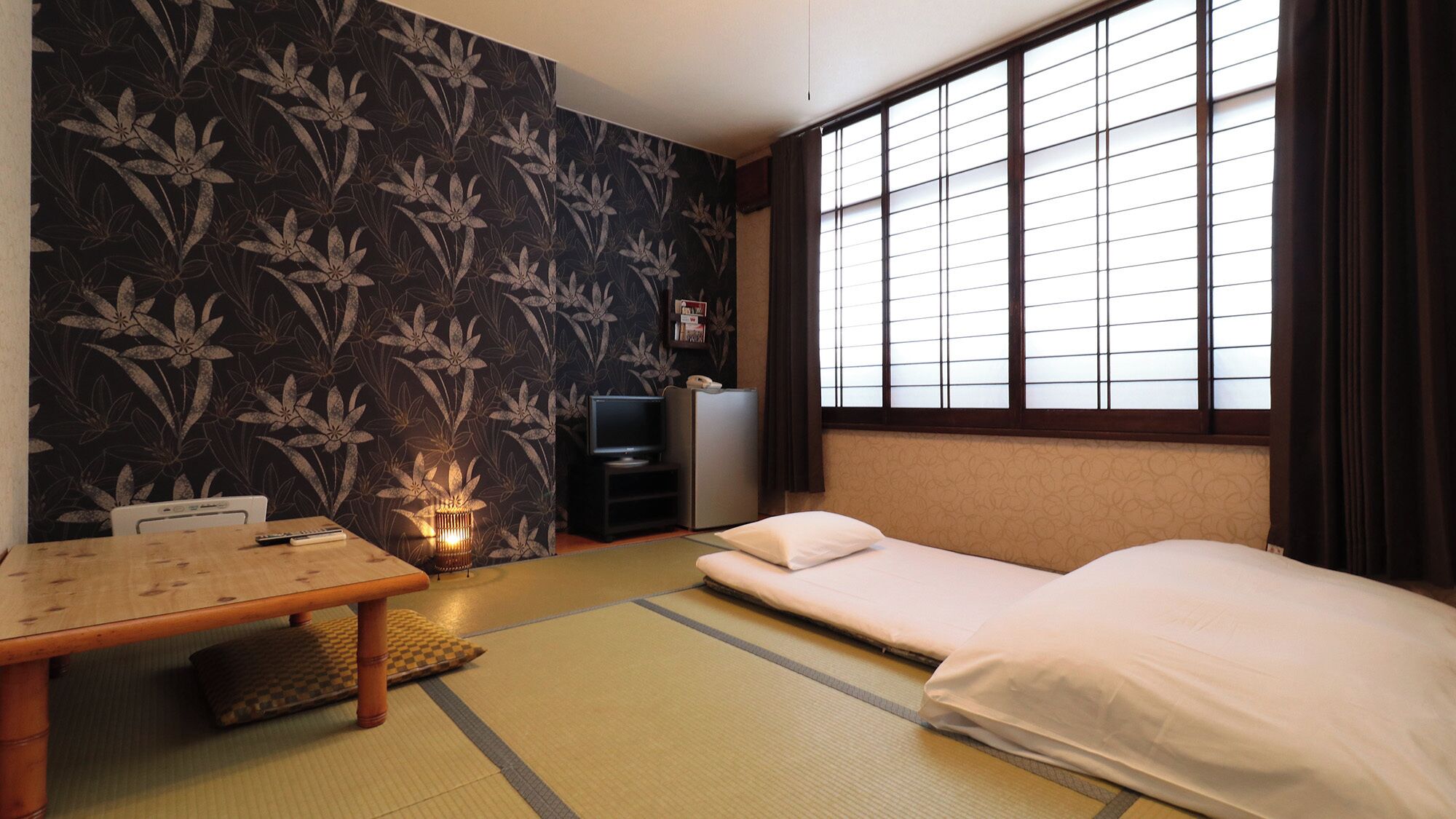 Ein traditioneller japanischer Raum mit Tatami-Böden, einem niedrigen Tisch, einem Bett, einem Fernseher und einem Kühlschrank.