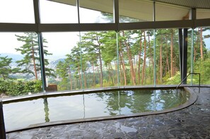 Une piscine intérieure au sol en pierre, dotée d’un mur en verre qui offre une vue sur de grands arbres et des montagnes lointaines.