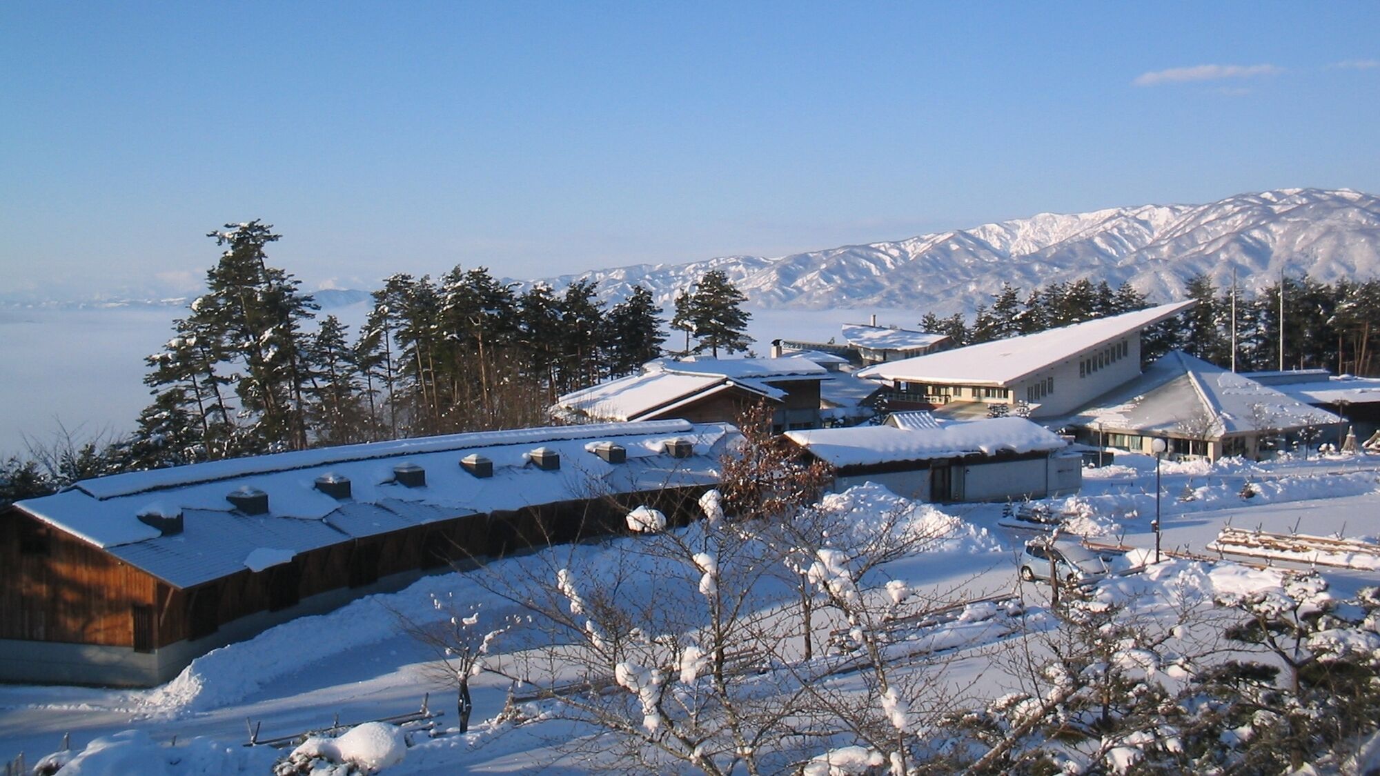 A snow-covered resort with multiple buildings, including a large lodge with a sloped roof, surrounded by trees and mountains in the background.