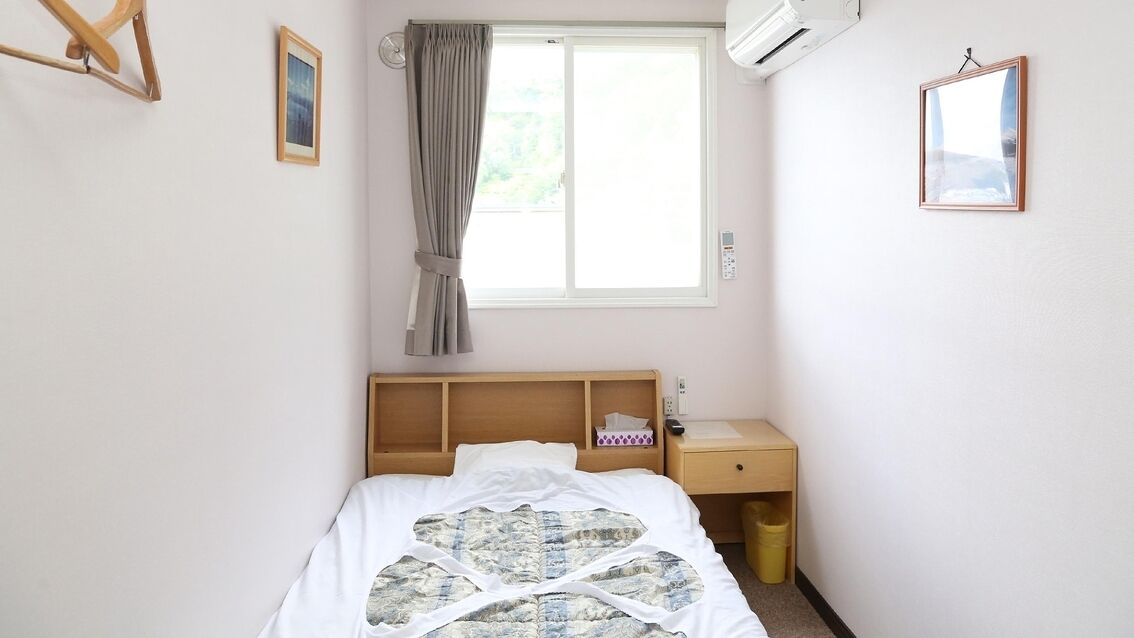 A bedroom with a bed, a bedside table, a nightstand, a window with curtains, and a wall-mounted air conditioner.