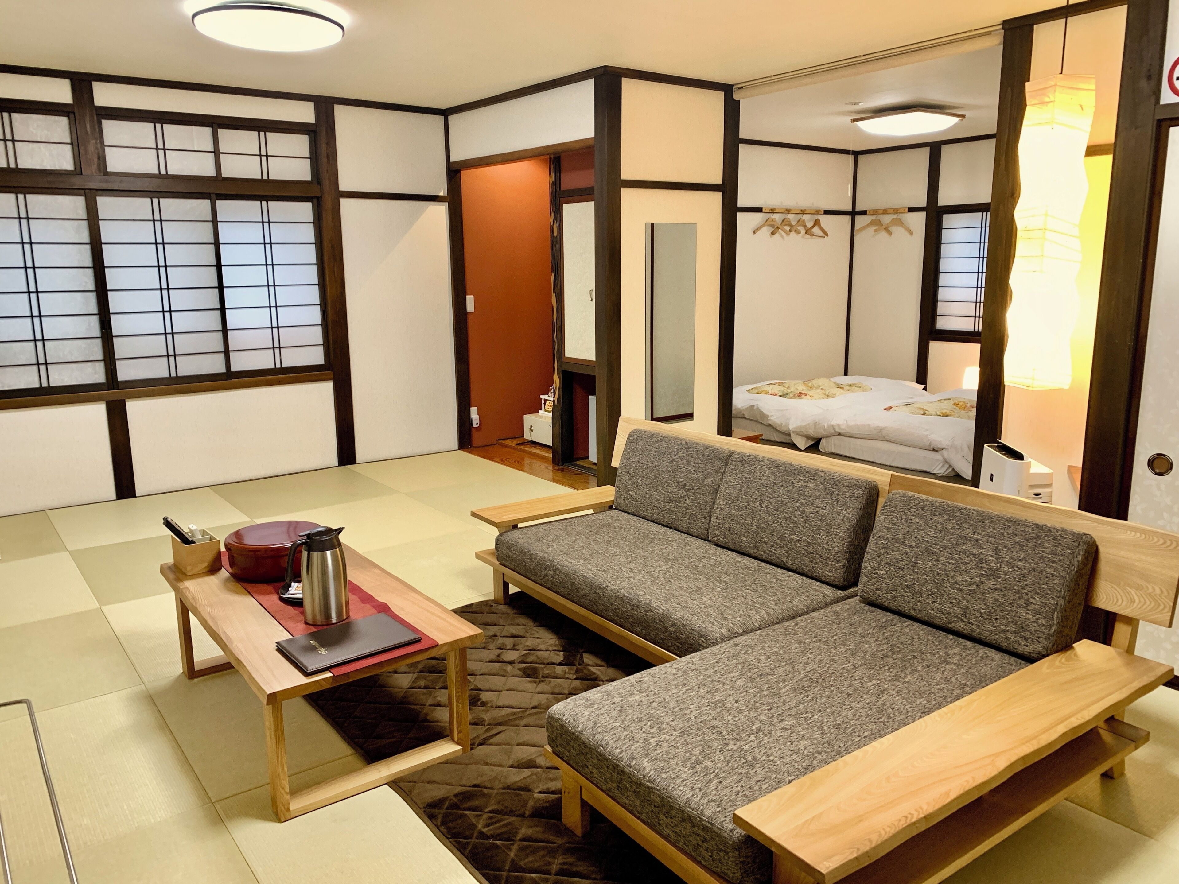 Une pièce de style japonais traditionnel, avec un sol recouvert de tatamis, un canapé, une table avec une bouilloire et un lit surmonté d’une lampe suspendue.