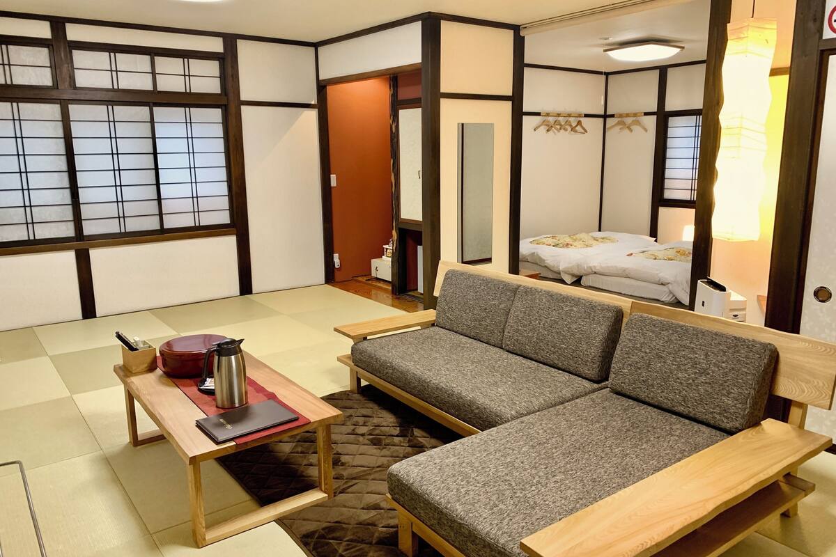 Um quarto em estilo japonês tradicional, com tatames, um sofá, uma mesa com chaleira e uma cama com abajur.