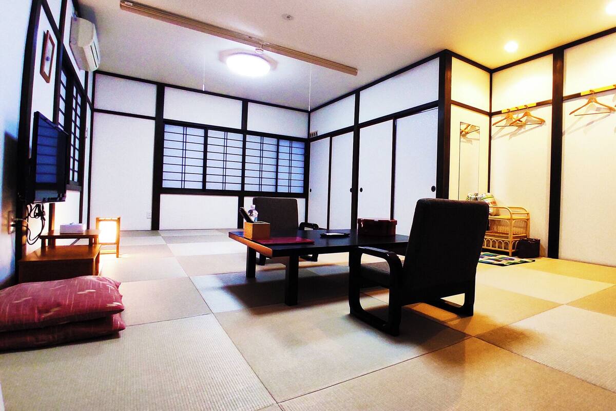 Um quarto japonês tradicional com tatames, uma mesa baixa, uma cadeira, um futon e um portas correderas com padrão quadriculado.