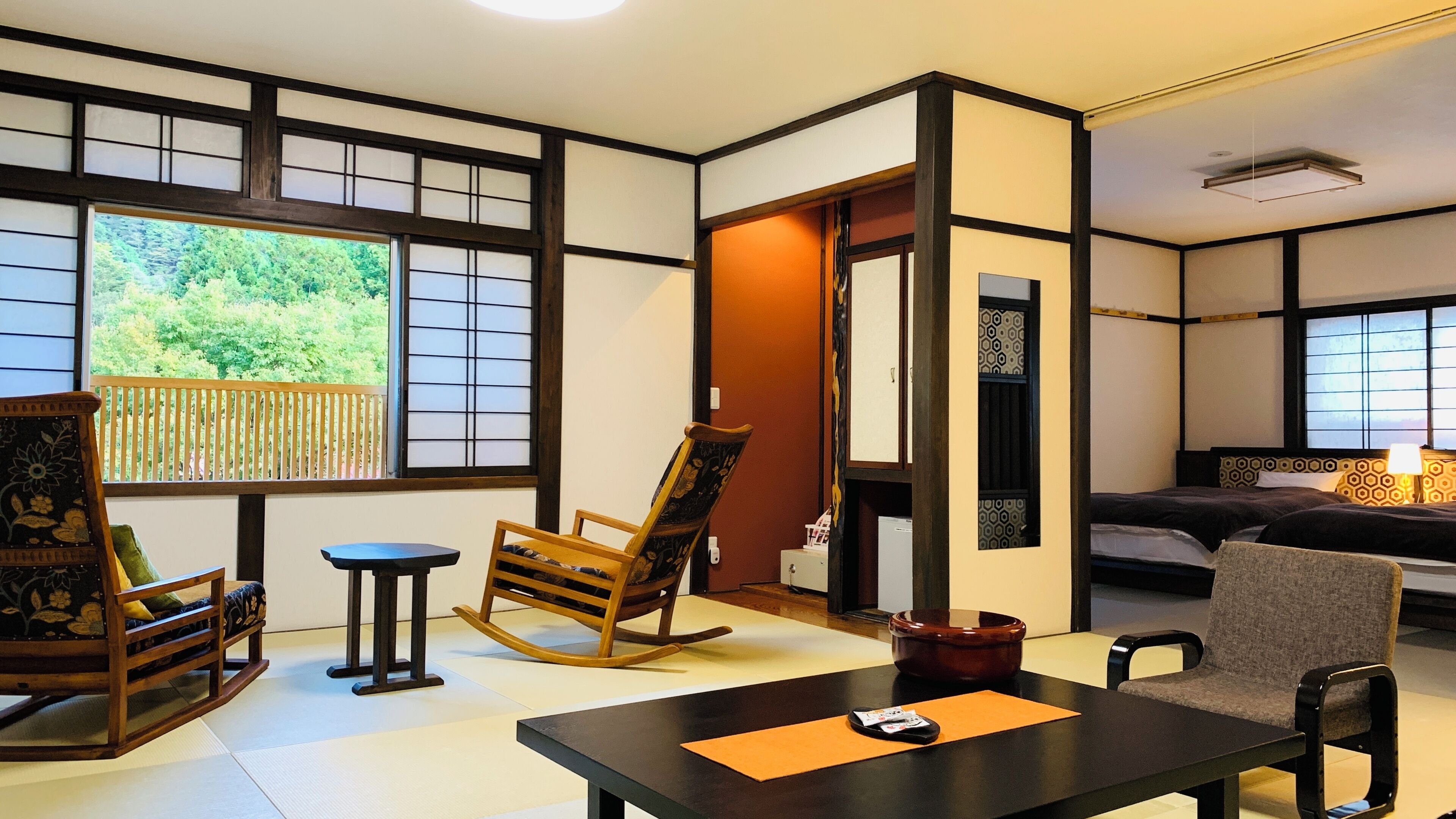Une pièce de style japonais traditionnel, avec un sol recouvert de tatamis, une table basse, un fauteuil à bascule, un canapé et un lit doté d’une tête de lit à motifs.