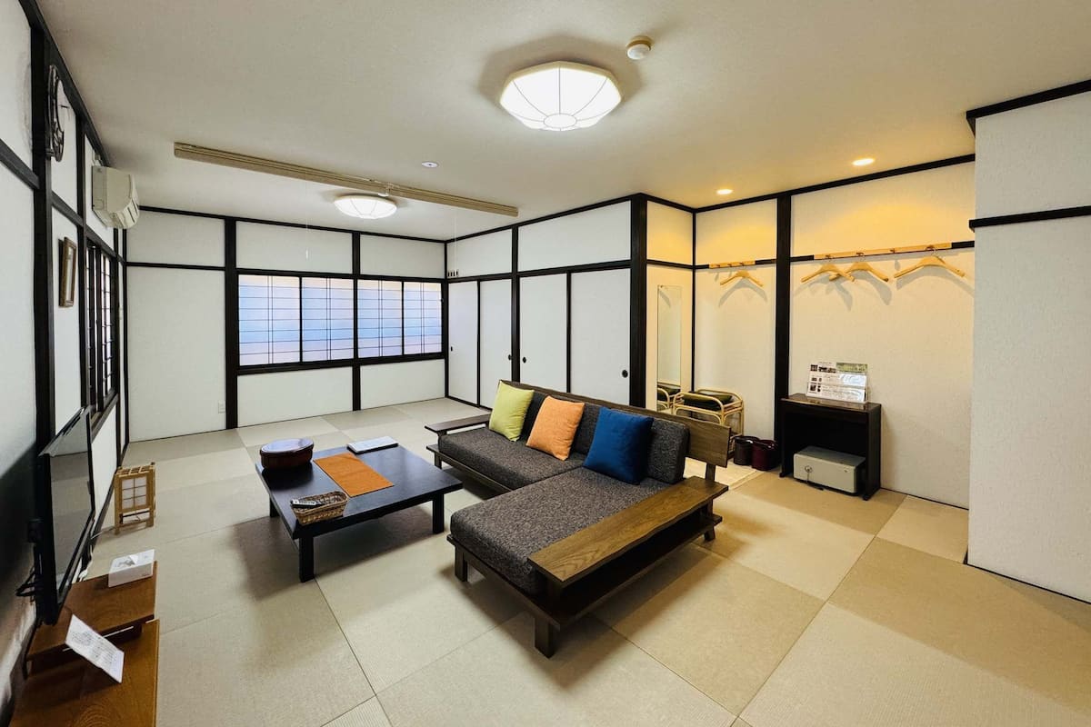 Quarto tradicional japonês com tatames, uma mesa baixa, um sofá com almofadas, uma pequena mesa com um cesto e uma prateleira com objetos.