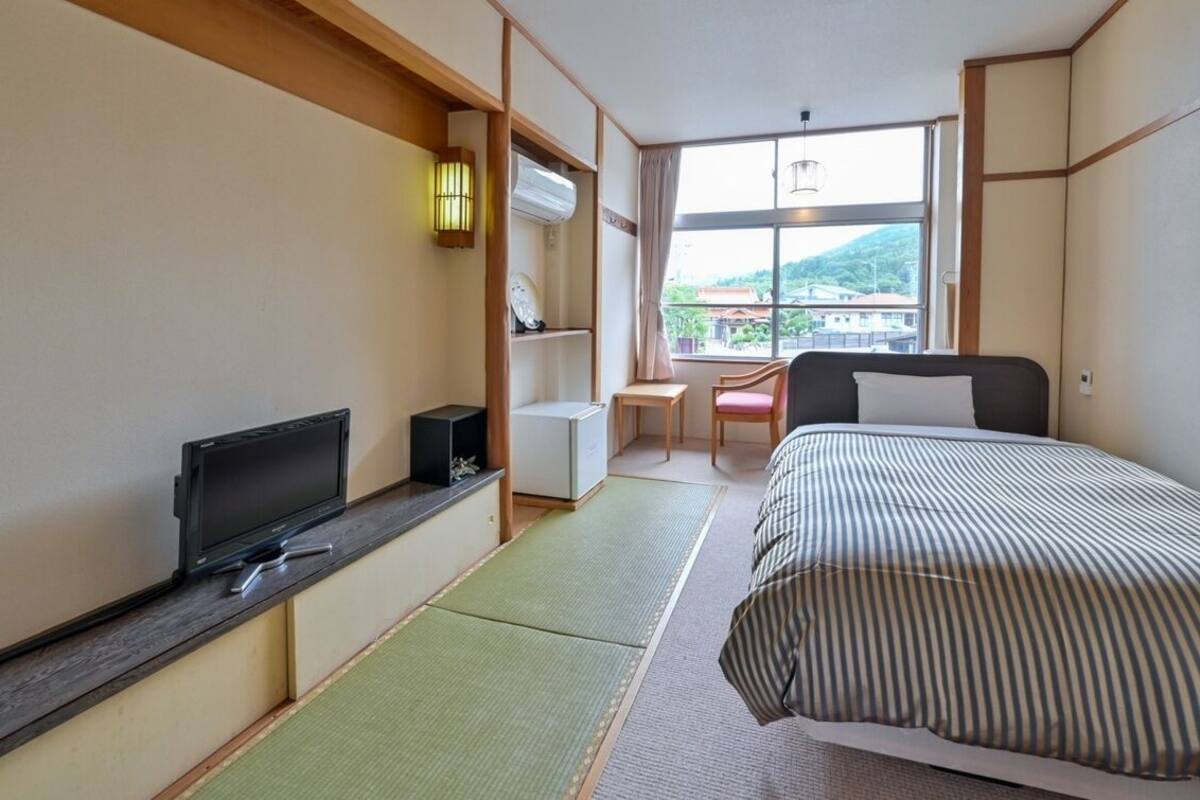 A hotel room with a bed, a desk with a chair, a television, and a window with a view of buildings and hills.