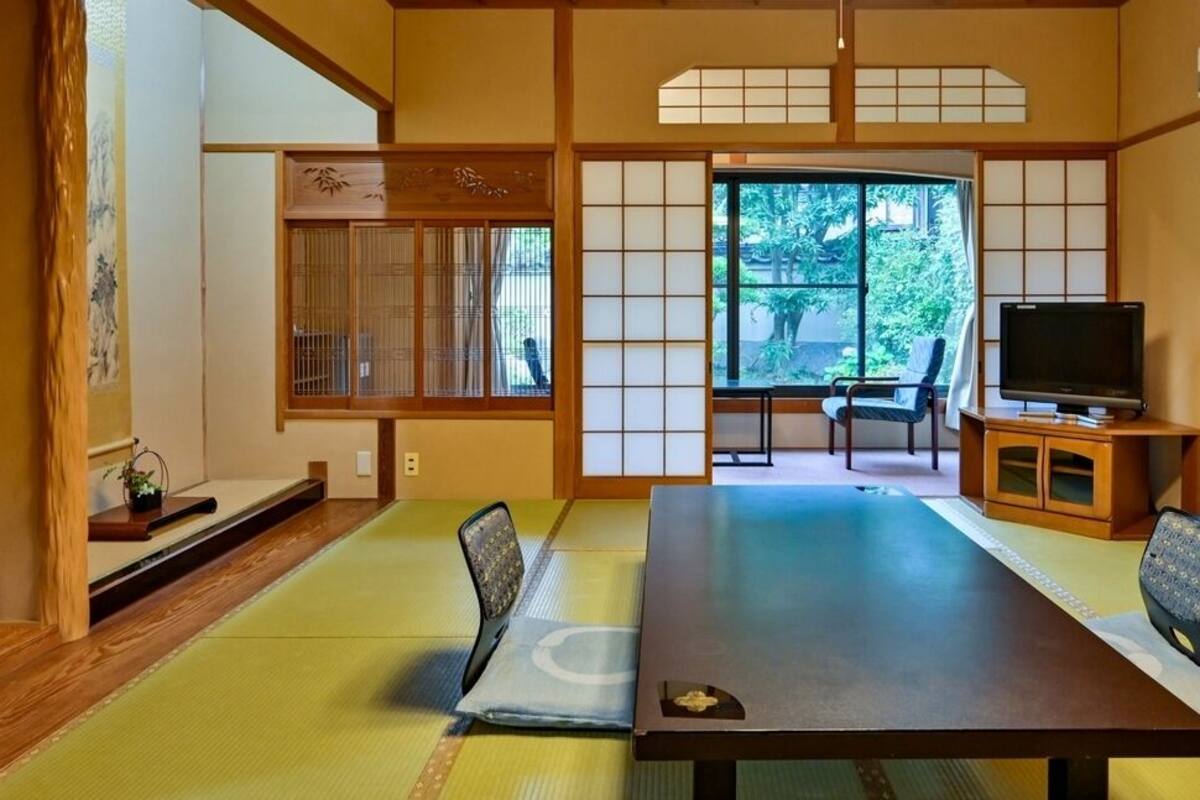 A traditional Japanese room with tatami flooring, a low table, chairs, a television, and a view of greenery outside.