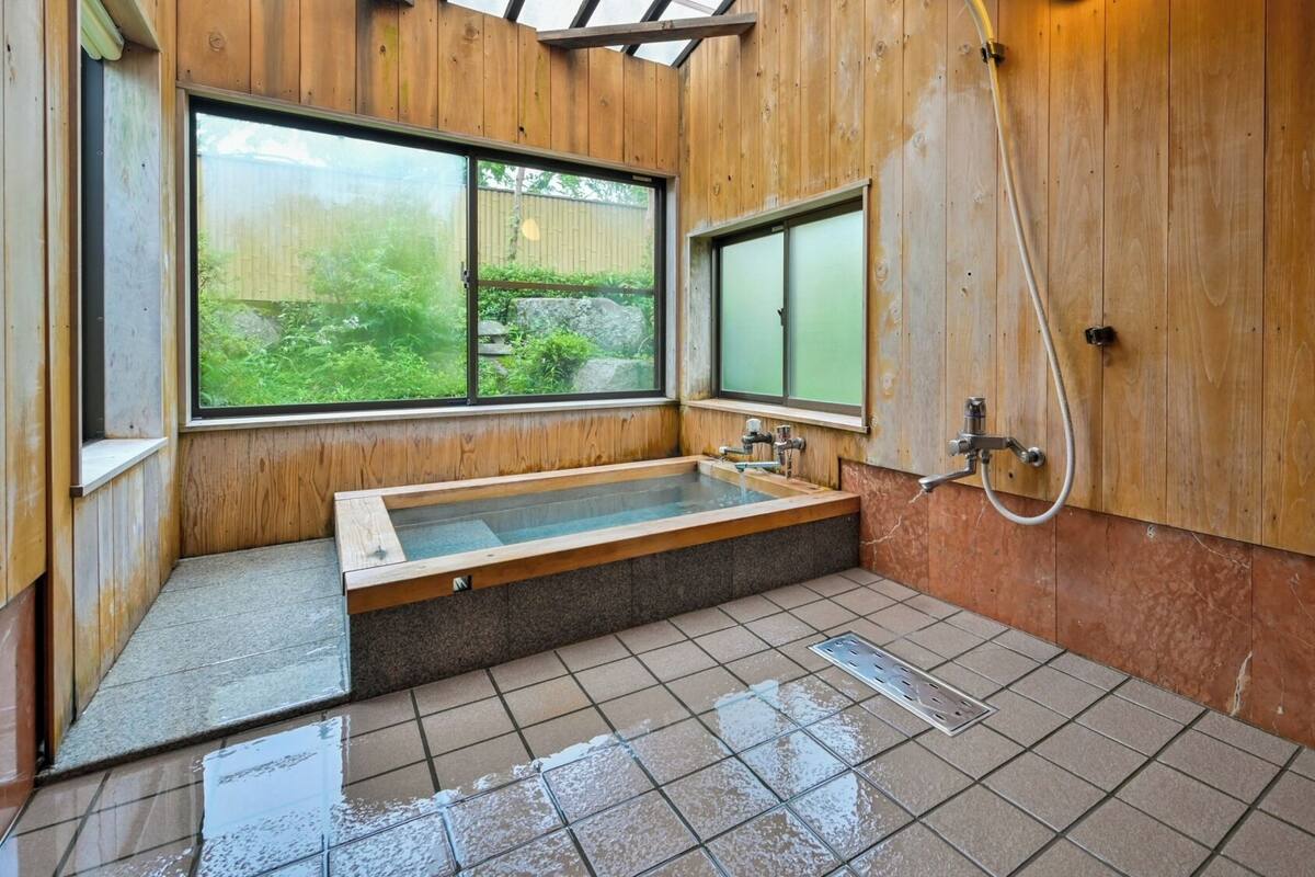 A wooden bathroom with a large bathtub, a shower, and a window providing a view of greenery.