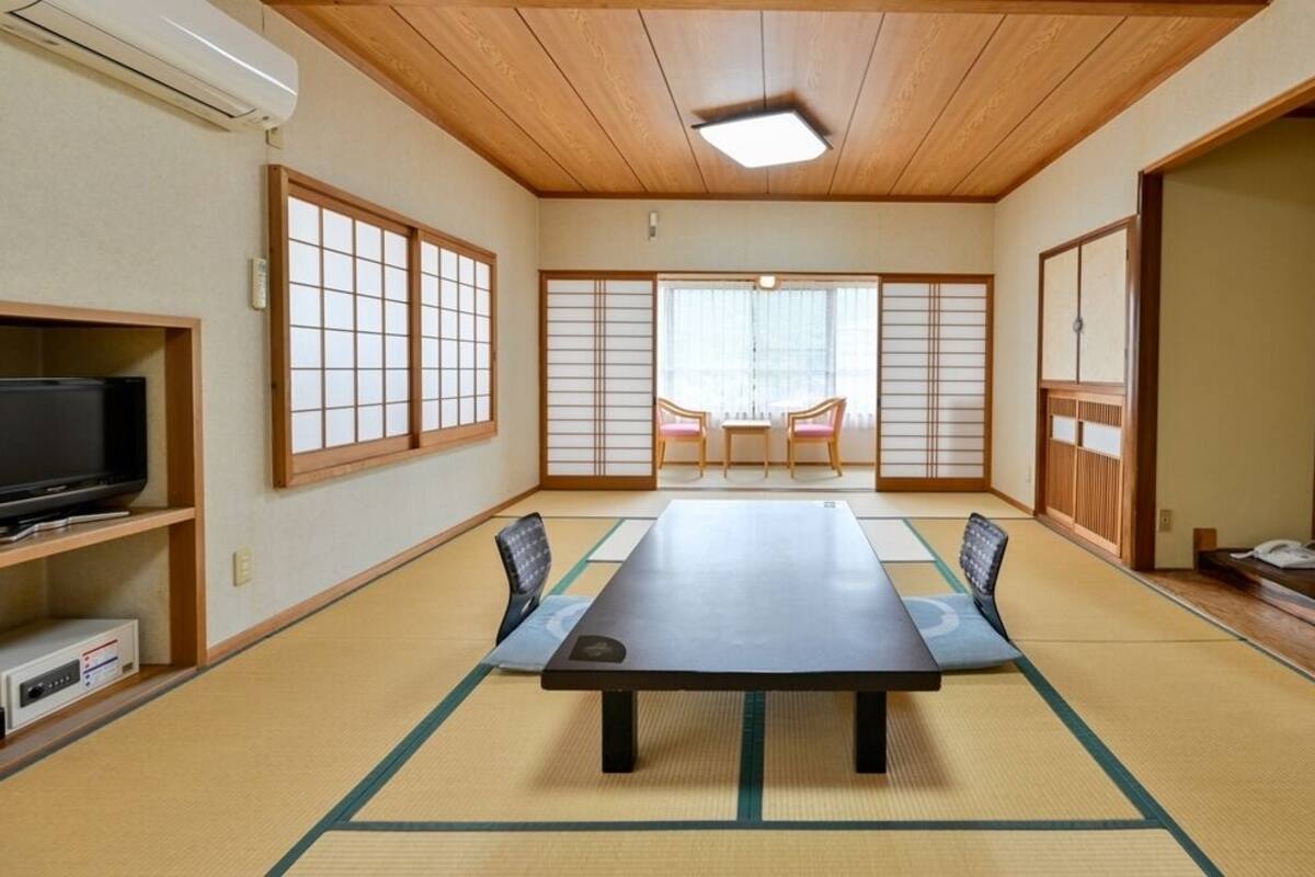 A traditional Japanese room with tatami flooring, a tatami mat table, a television on the left, sliding doors, and a view through the windows.