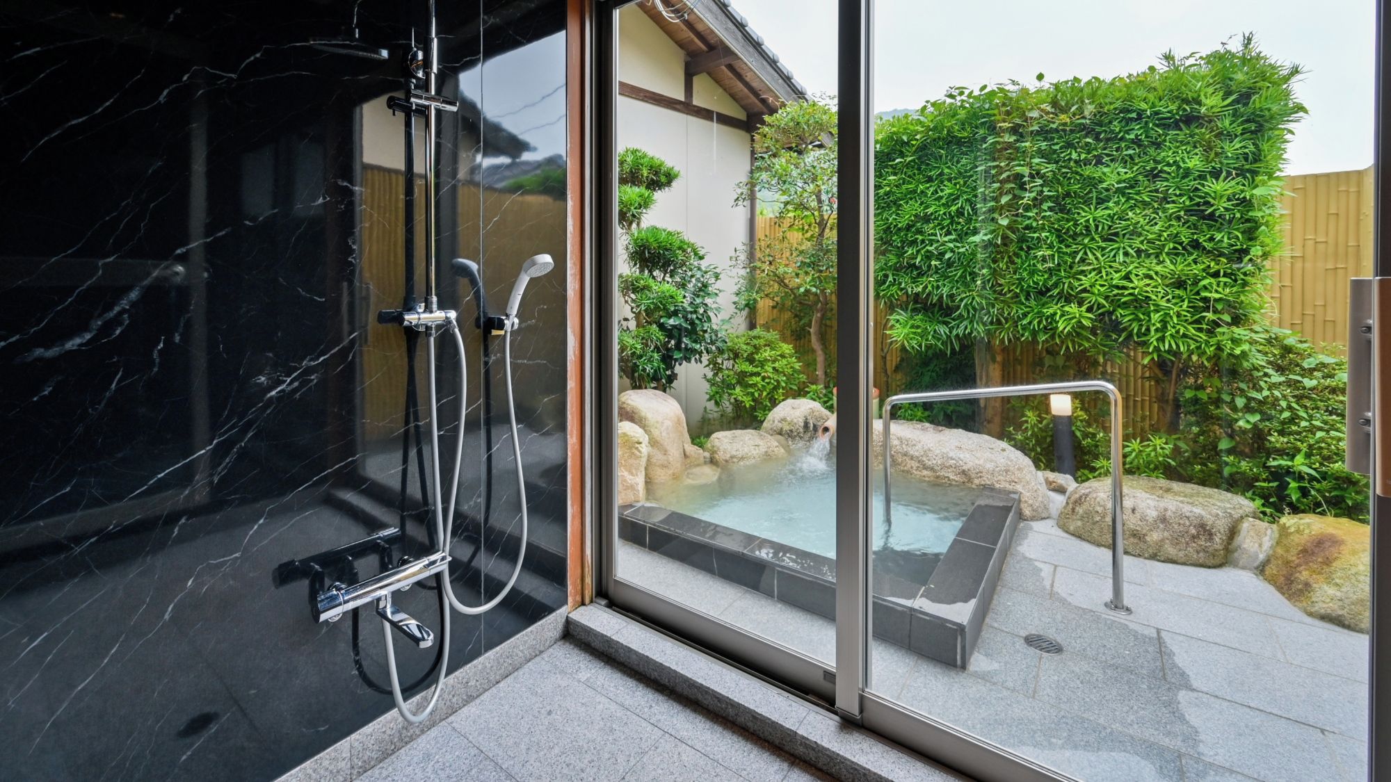 A modern bathroom with a glass shower enclosure, a hot tub, and a view of a landscaped garden.