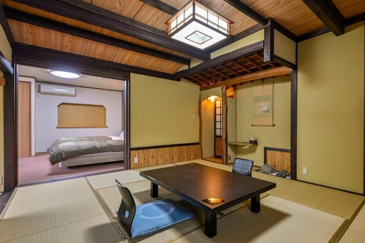 A traditional Japanese room with tatami flooring, a low table, a chair, a bed in the background, and a wooden ceiling with a skylight.