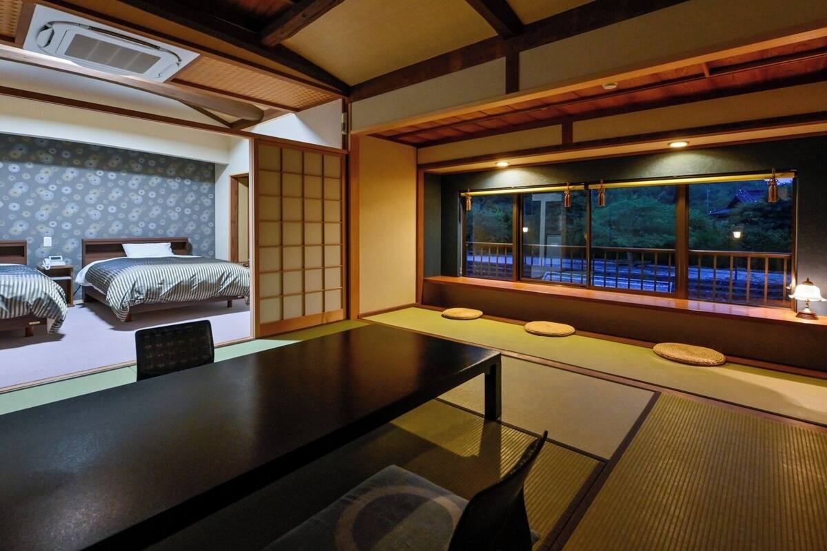 A traditional Japanese-style room with tatami flooring, a low table, two beds with striped bedding, and a view of a garden through a window.