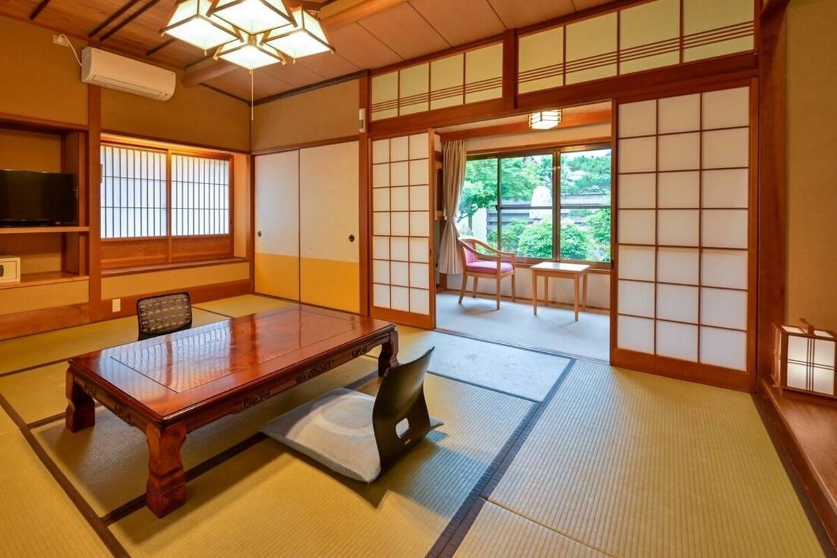 A traditional Japanese room with tatami flooring, a wooden table, a television, sliding doors, and a view of greenery outside.