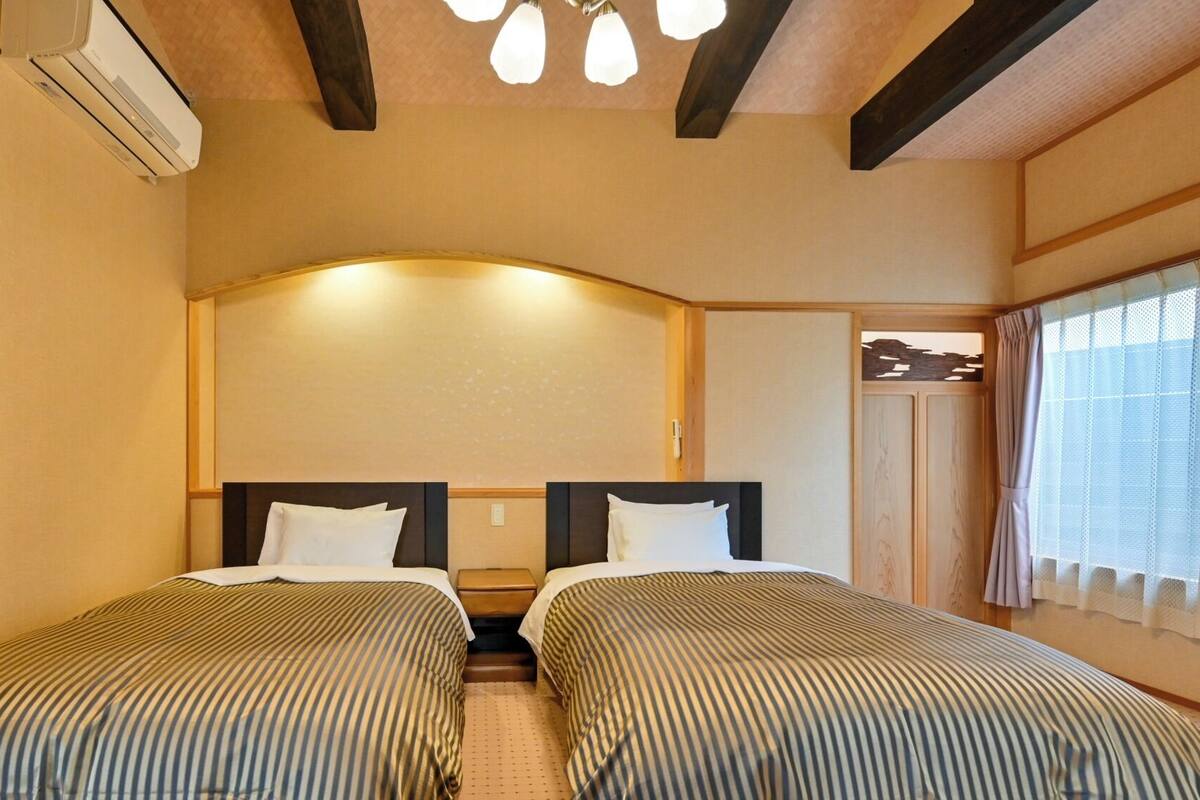 A hotel room with two beds, a wooden ceiling, an air conditioning unit, and a window with curtains.