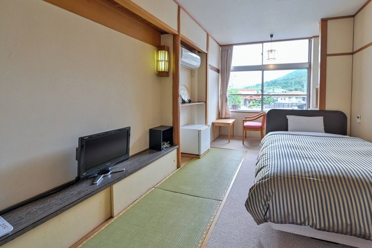 A hotel room with a bed, a television, a desk, and a window with a view of buildings.