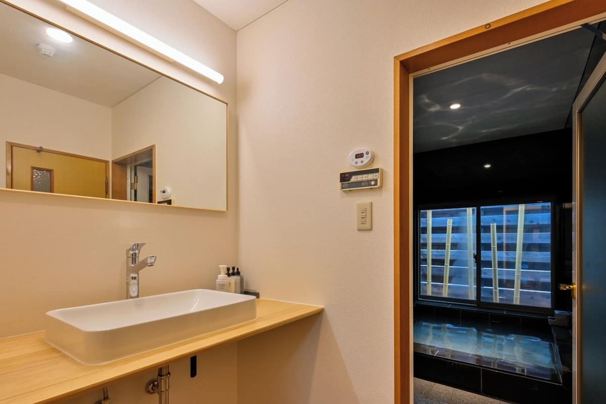 A bathroom with a large mirror, a white sink, a handheld soap dispenser, and a digital thermostat on the wall.