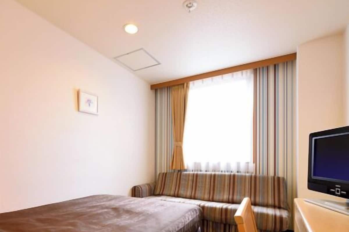 Une chambre d’hôtel avec un lit, un bureau avec un ordinateur, une fenêtre avec des rideaux et un petit tableau accroché au mur.