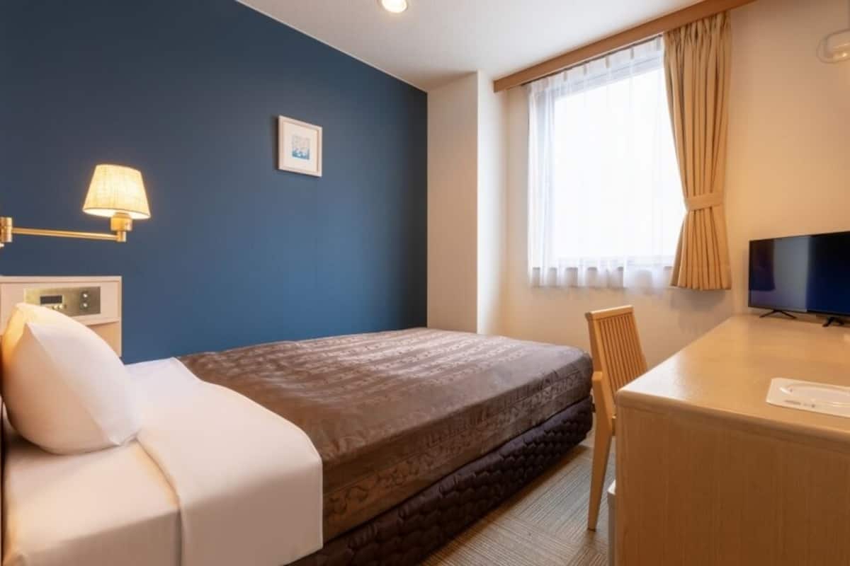 Une chambre d’hôtel avec un lit, un bureau, une télévision, une lampe et une fenêtre avec des rideaux.