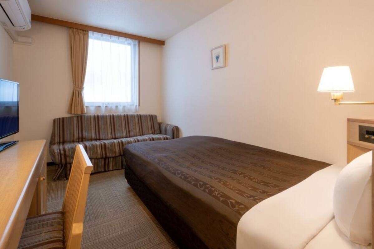 Une chambre d’hôtel avec un lit, un canapé, une télévision, une lampe et une fenêtre avec des rideaux.