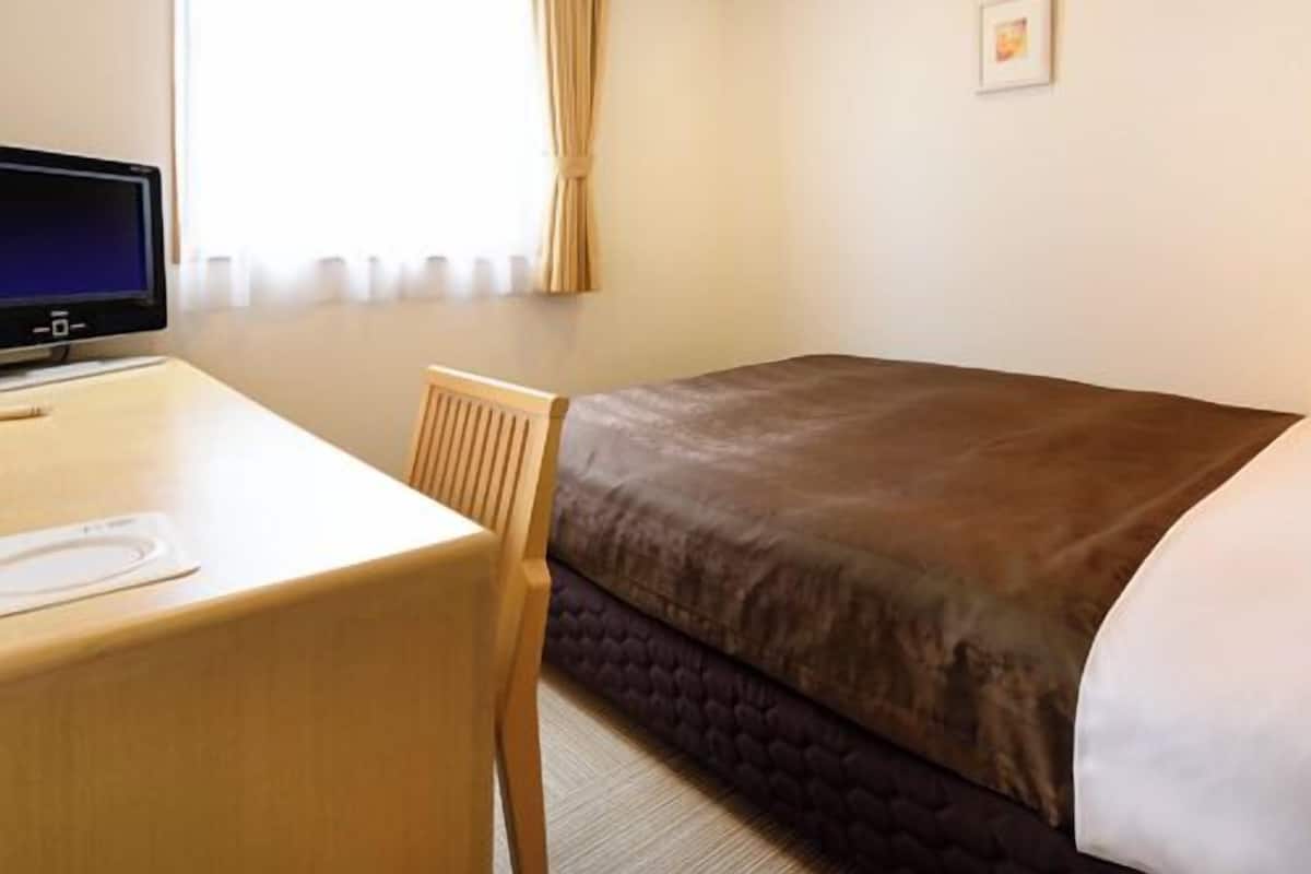 Une chambre d’hôtel avec un lit, un bureau, une chaise, une fenêtre avec des rideaux et un petit tableau au mur.