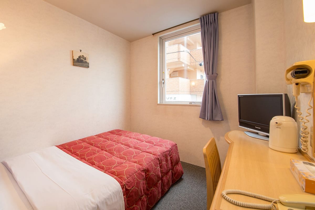A small hotel room with a single bed, a desk with a computer, a telephone, and a window with a curtain.