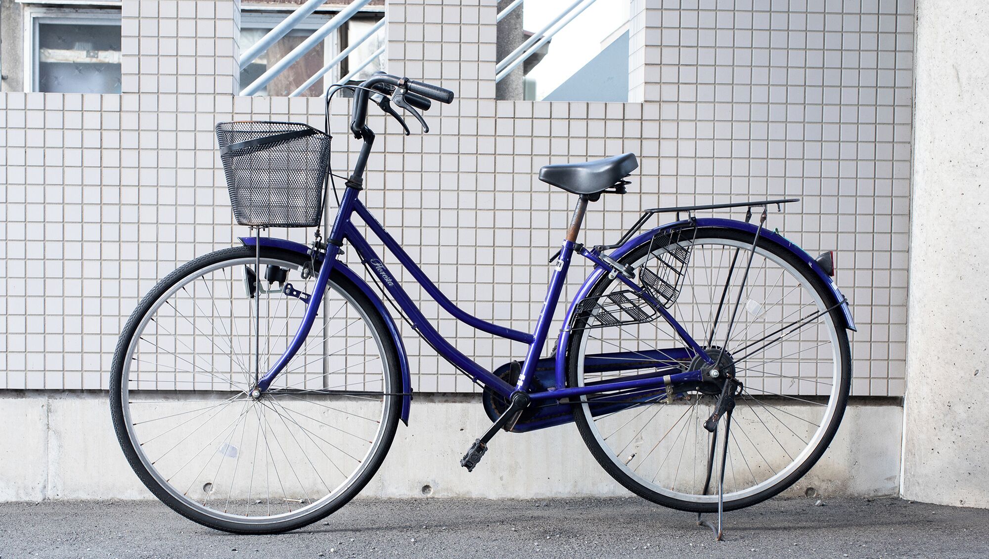 A blue bicycle with a basket and a seat, parked against a tiled wall.