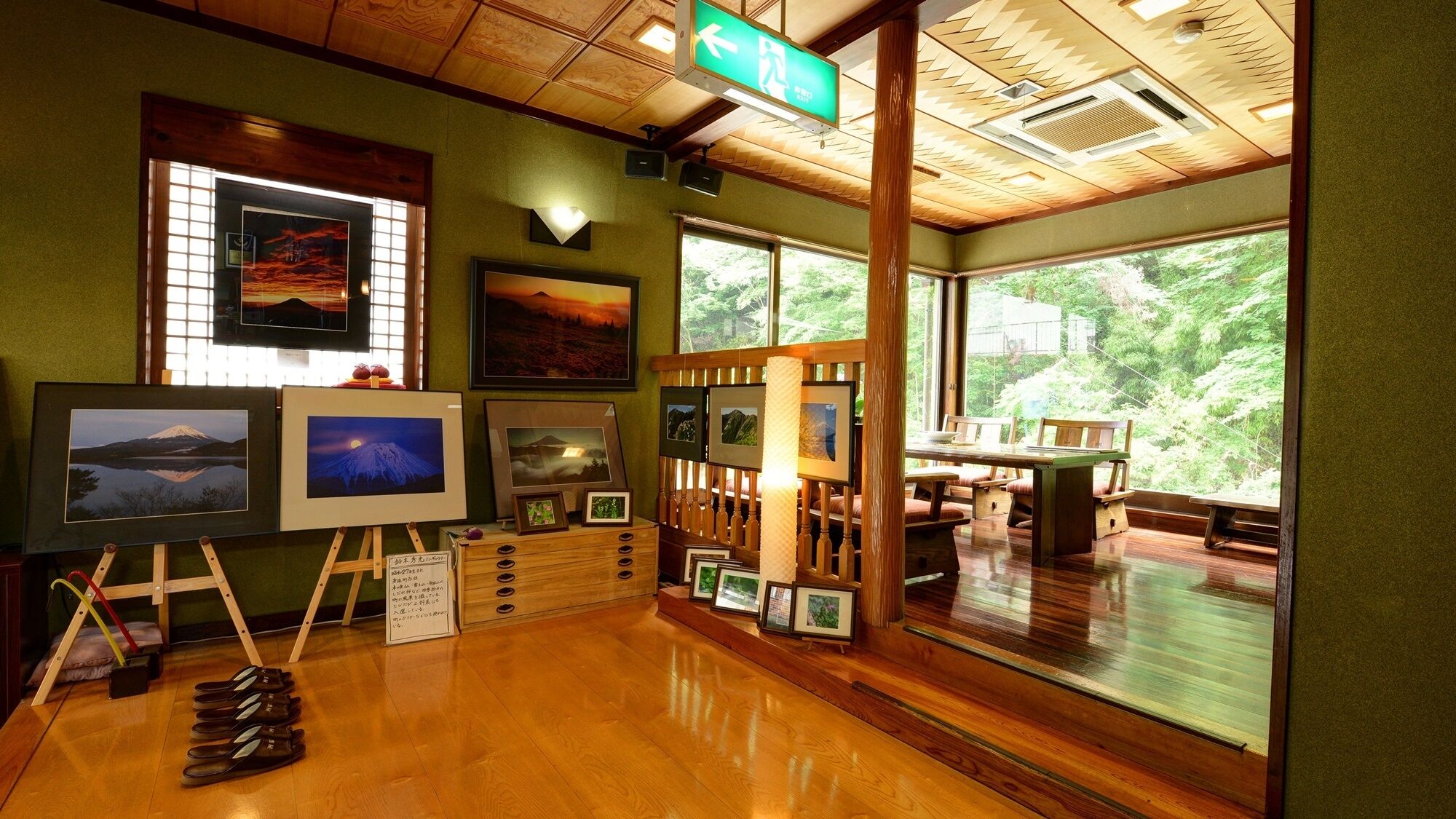 A room with wooden flooring, framed pictures on easels, a dining table, and large windows with a view of greenery.