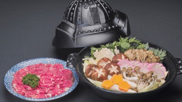 A traditional Japanese hot pot setup with a metal lid, a blue ceramic dish with pink-colored food, and a black pan with various vegetables and mushrooms.