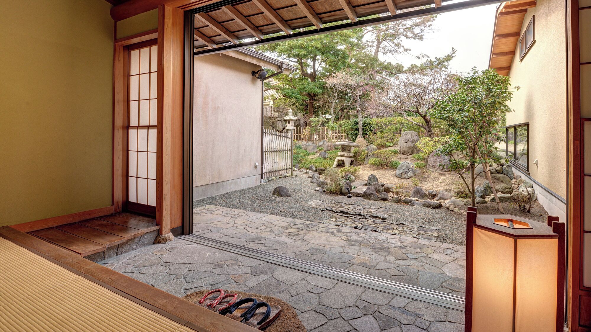 Un interior japonés tradicional con suelo de tatami, puertas correderas y vistas a un jardín ajardinado con rocas y un árbol pequeño.