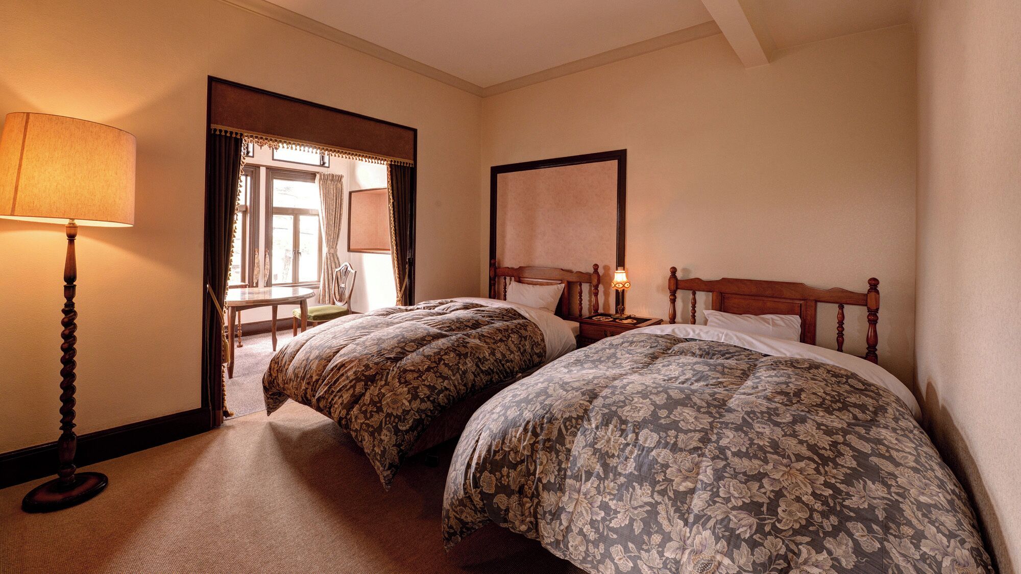 Una habitación de hotel con dos camas, un rincón de estar con una mesa y una silla, y un ventanal grande.