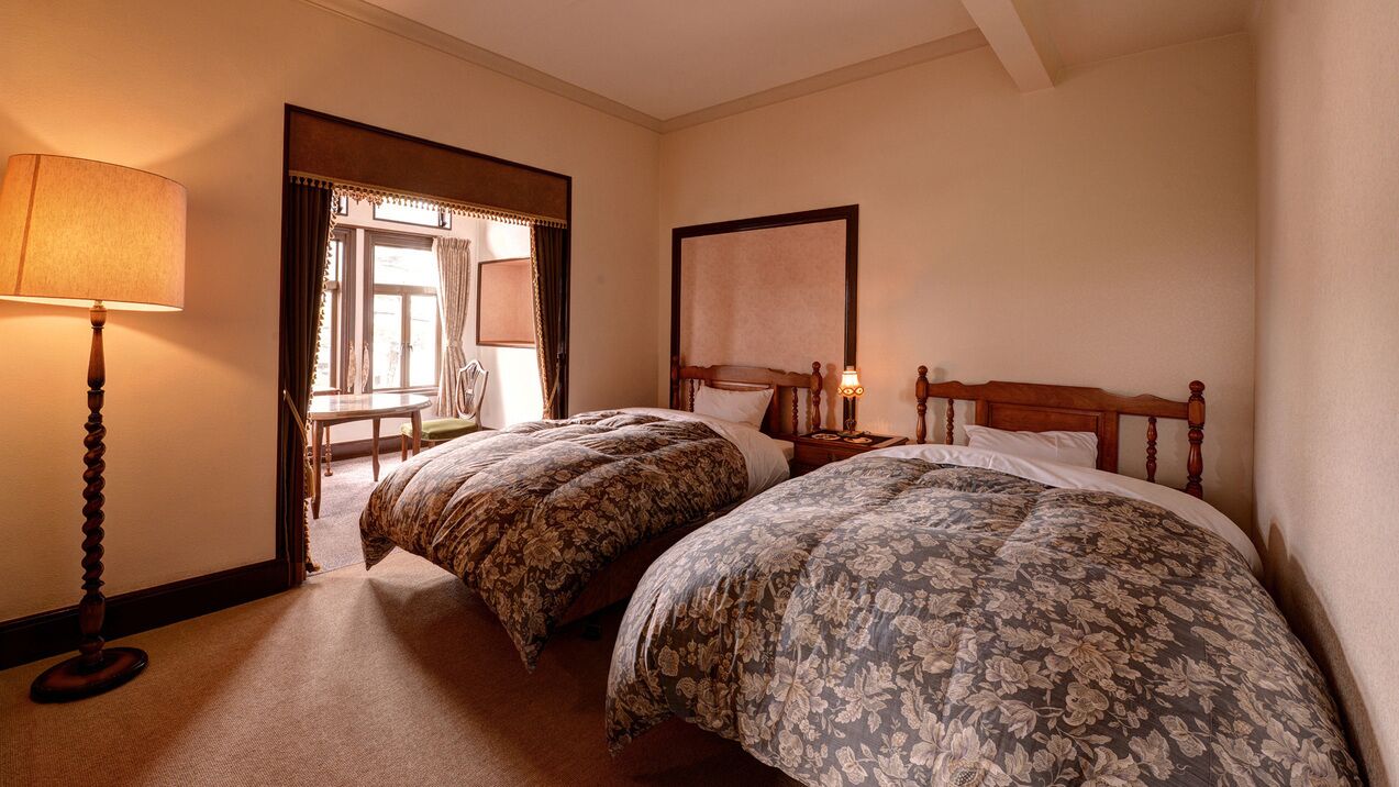Habitación de hotel con dos camas, una mesita de noche con lámpara, una silla y una ventana con cortinas.