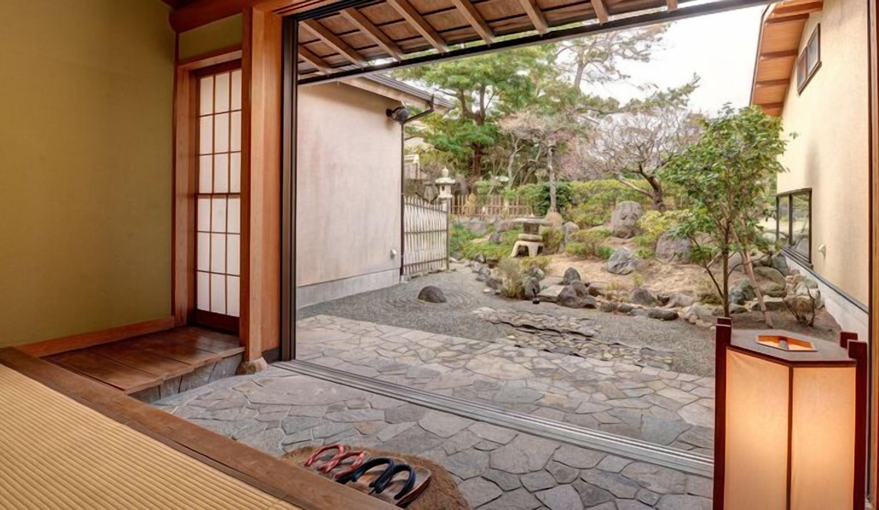 Una habitación japonesa tradicional con suelo de tatami, puertas correderas y vistas a un jardín paisajístico con rocas y árboles.