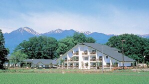 A multi-story building with a sloped roof, surrounded by a green field and trees, with a mountain range in the background. - Village Azumino (Azumino)