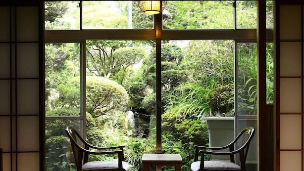 A room with two chairs and a table, large windows with a view of greenery and a stone structure.