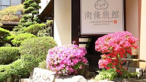 A traditional Japanese building with a signboard, surrounded by well-maintained bonsai trees and flowering shrubs.