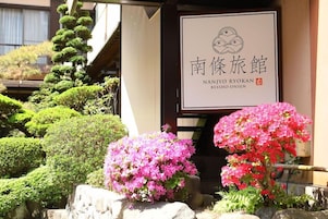 Un edificio japonés tradicional con un cartel, rodeado de bonsáis bien cuidados y arbustos florecientes.