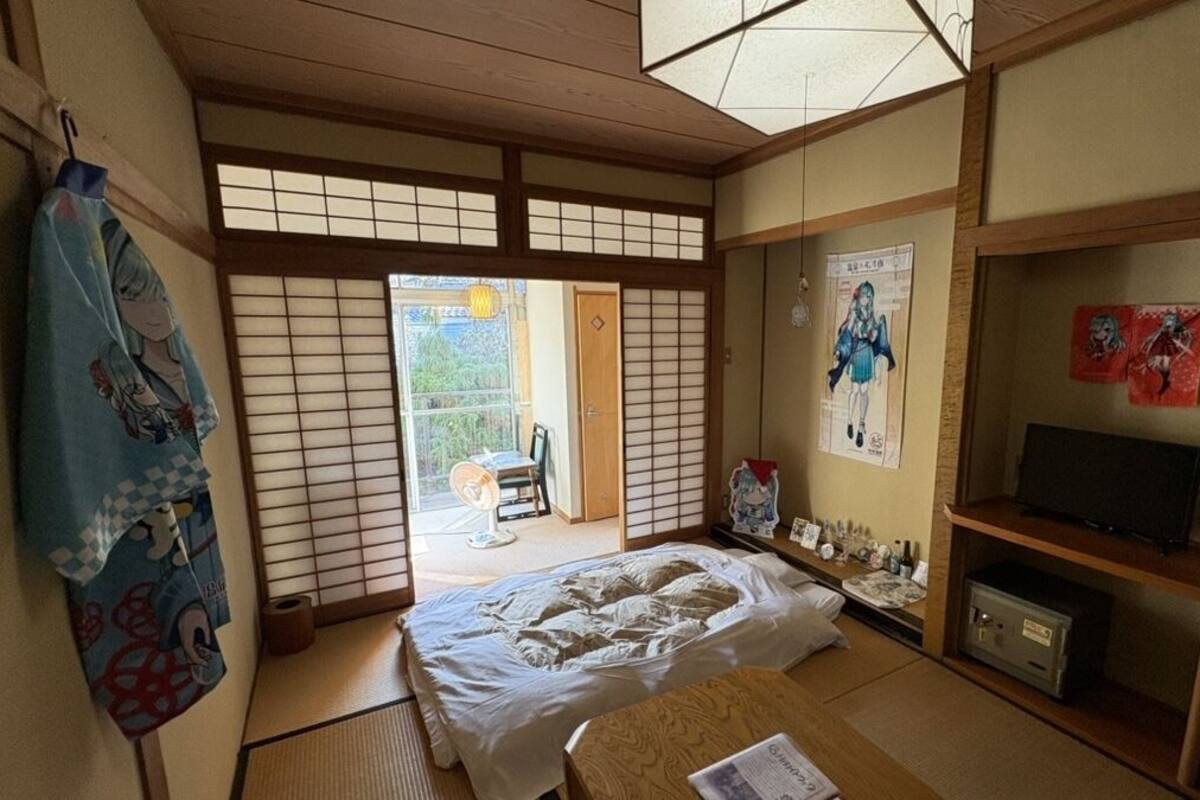Una habitación japonesa tradicional con suelo de tatami, una cama futón, un televisor y una puerta corredera que da a un espacio exterior.