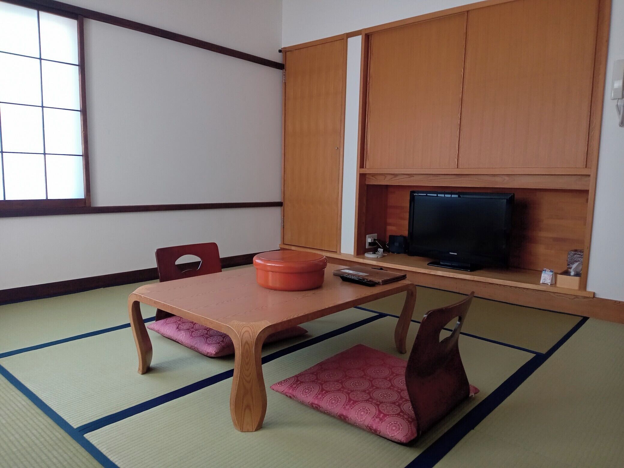 A traditional Japanese room with tatami flooring, a low table, two wooden chairs, a television on a shelf, and a sliding door.