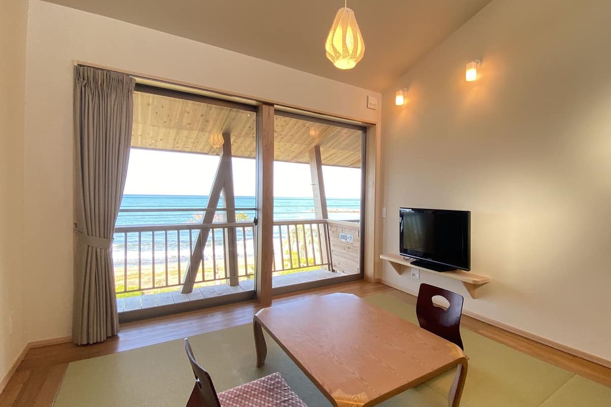 A room with a TV, a table, a chair, and a view of the ocean through large sliding doors.