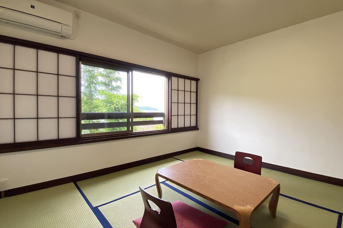 A room with tatami flooring, a low table, a chair, and a shoji screen with a view of greenery outside.