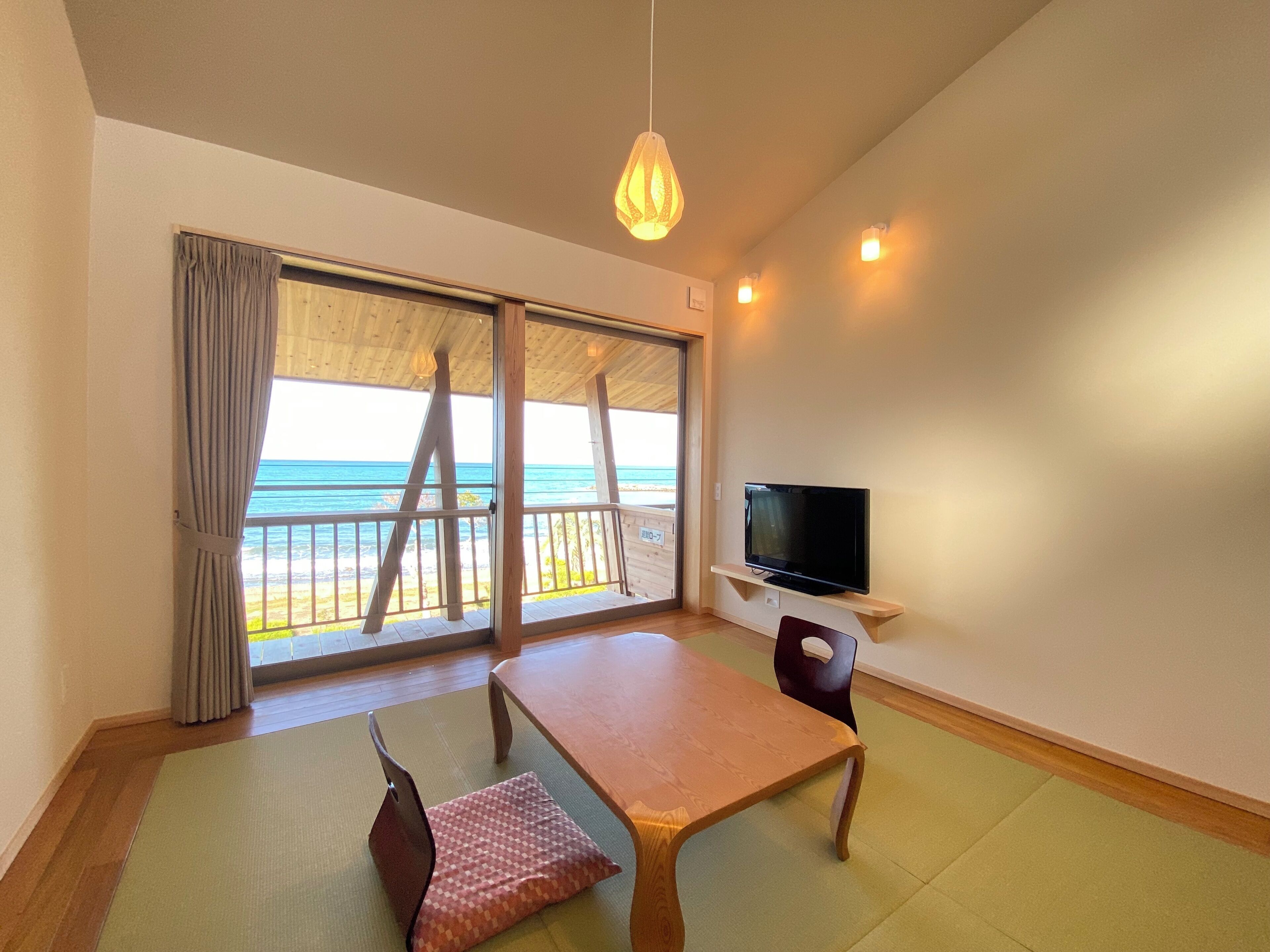 A room with a wooden table, a TV on a shelf, a chair, a view of the beach, and a large window with curtains.