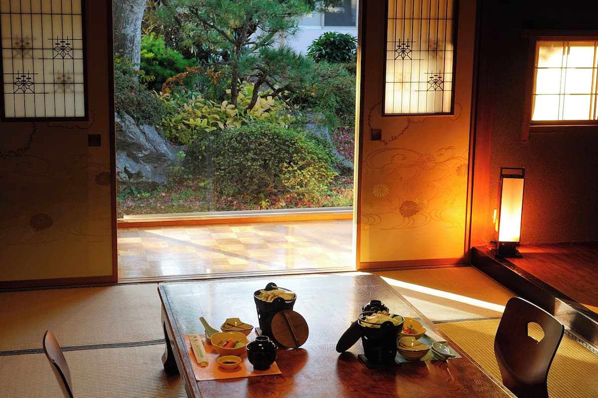 A traditional Japanese room with tatami flooring, a low wooden table with tea set, and a view of a garden through shoji screens.