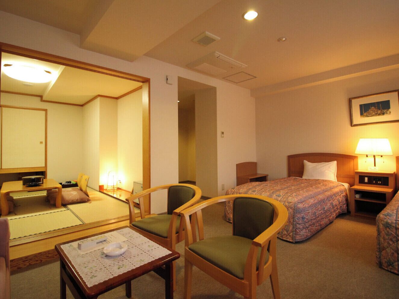 A hotel room with a bed, two chairs, a small table, a bedside table with a lamp, and a mirror.