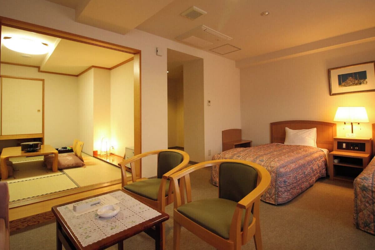 A hotel room with a bed, two chairs, a small table, a bedside table with a lamp, and a mirror.