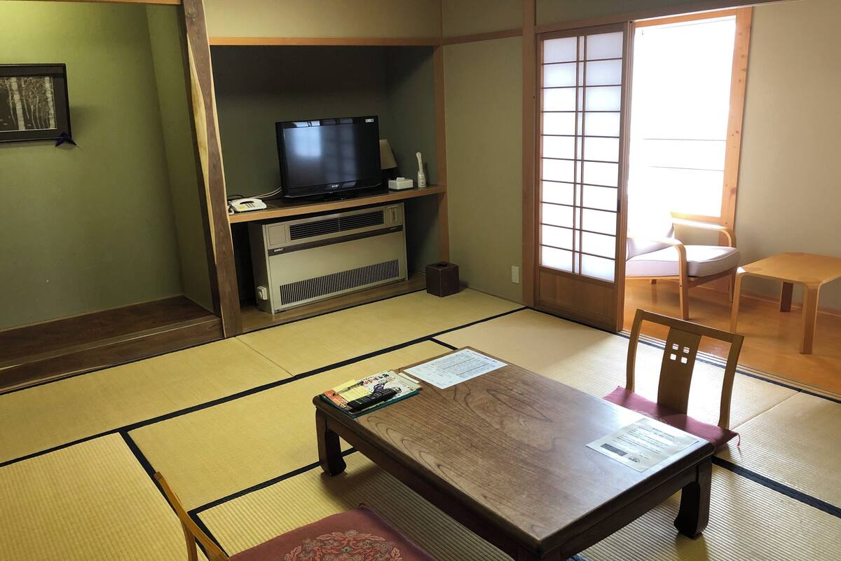 Ein traditioneller japanischer Raum mit Tatami-Böden, einem Fernseher auf einem niedrigen Tisch, einem Holztisch und einer verschiebbaren Tür.